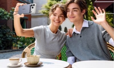 A smiling young man and woman sitting at an outdoor cafe, with the woman taking a selfie using an iPhone attached to a UGREEN magnetic power bank while the man waves at the camera.
