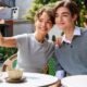 A smiling young man and woman sitting at an outdoor cafe, with the woman taking a selfie using an iPhone attached to a UGREEN magnetic power bank while the man waves at the camera.