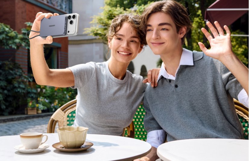 A smiling young man and woman sitting at an outdoor cafe, with the woman taking a selfie using an iPhone attached to a UGREEN magnetic power bank while the man waves at the camera.
