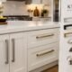 Realistic kitchen interior featuring stylish and durable KT&CO kitchen handles in brushed nickel, matte black, and brass finishes on cream cabinetry.